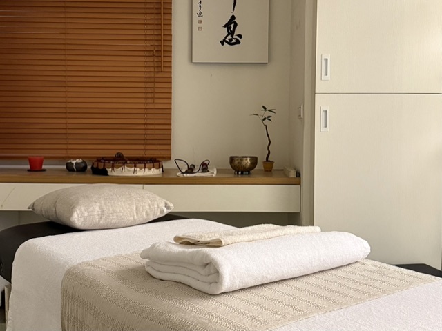 Inside the studio: a sheet-covered table by wooden blinds, a singing bowl and a hanging scroll on the shelf